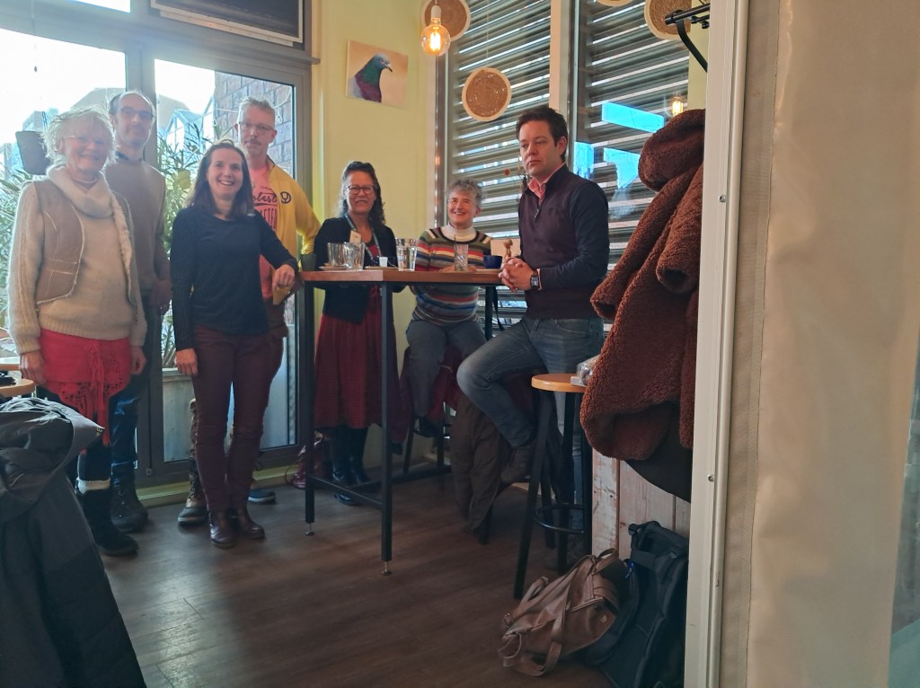 Groep mensen staat en zit in het gezellige Bakkerscafé, zichtbaar blij en betrokken, tijdens Open Coffee Nijmegen.